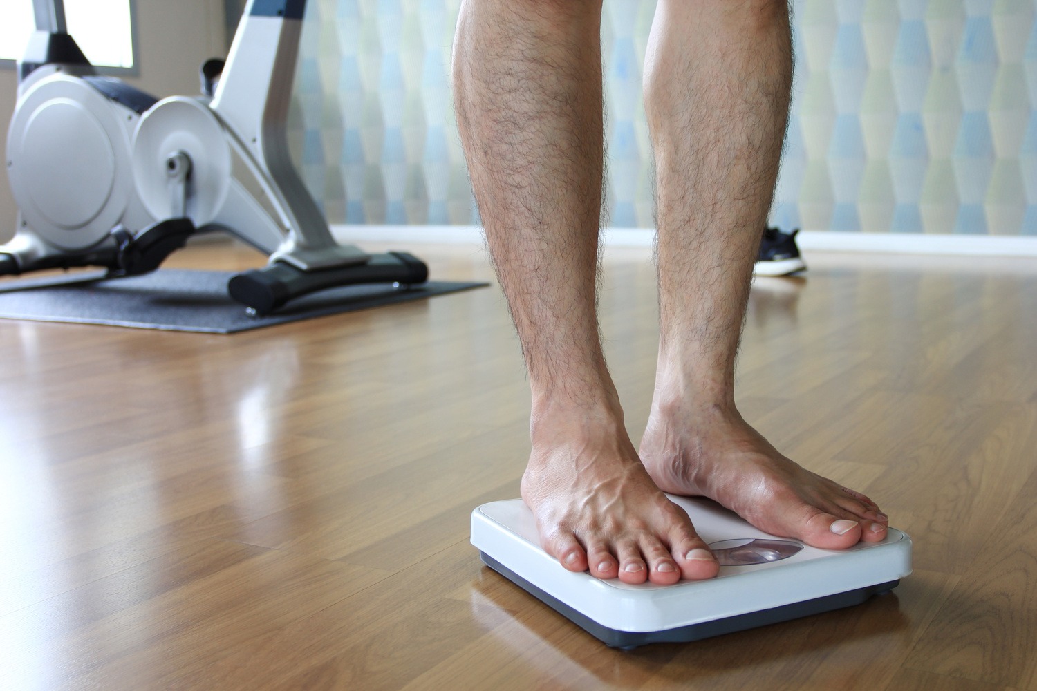 Bare feet standing on a digital scale on a wooden floor with exercise equipment in the background.