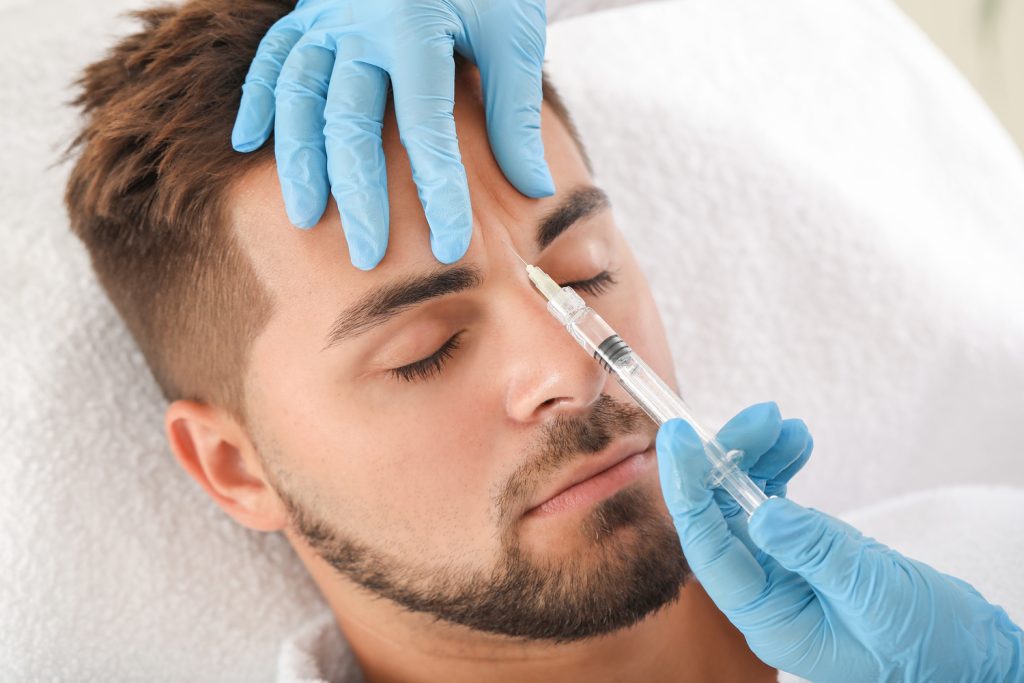 Gloved hands hold a syringe near a man’s forehead as he lies on a treatment bed with eyes closed