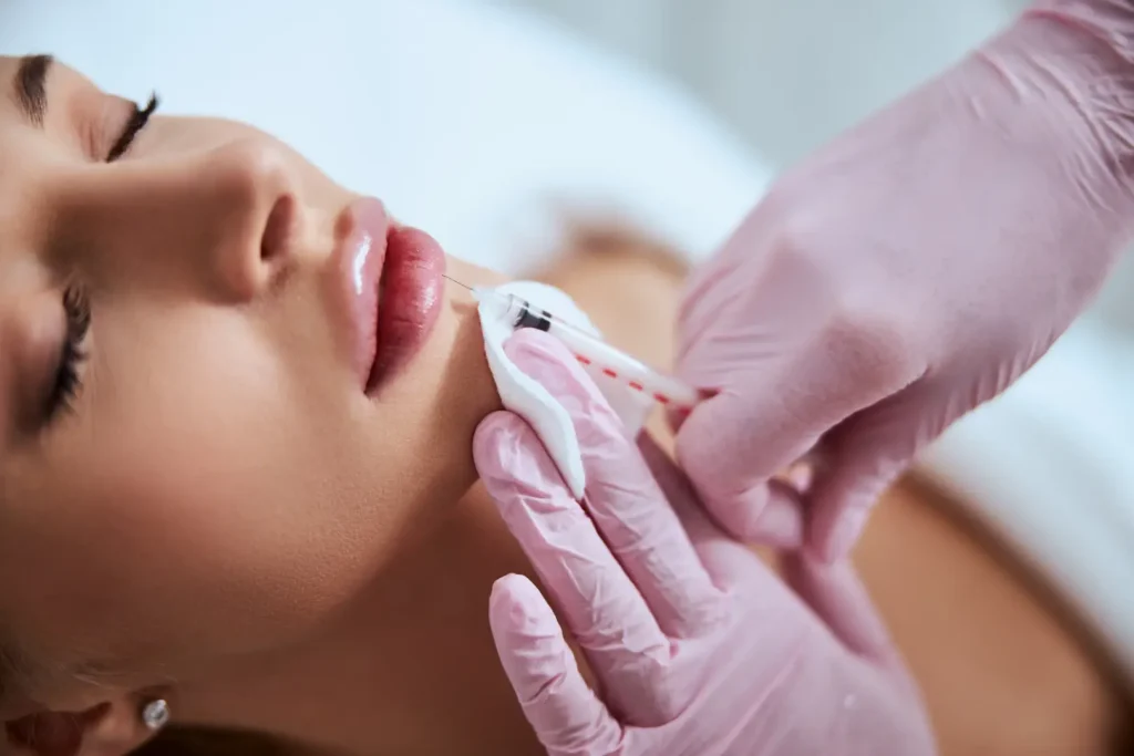Gloved hands holding a syringe near a woman’s lips during a cosmetic procedure