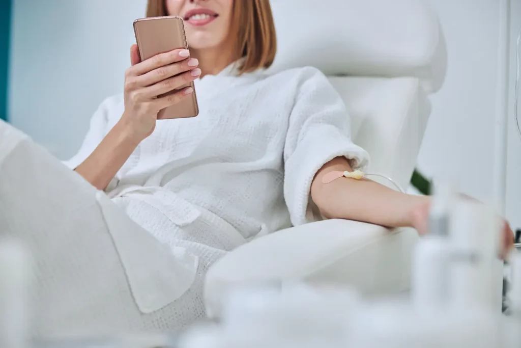 Woman in a white robe using a phone while receiving IV therapy