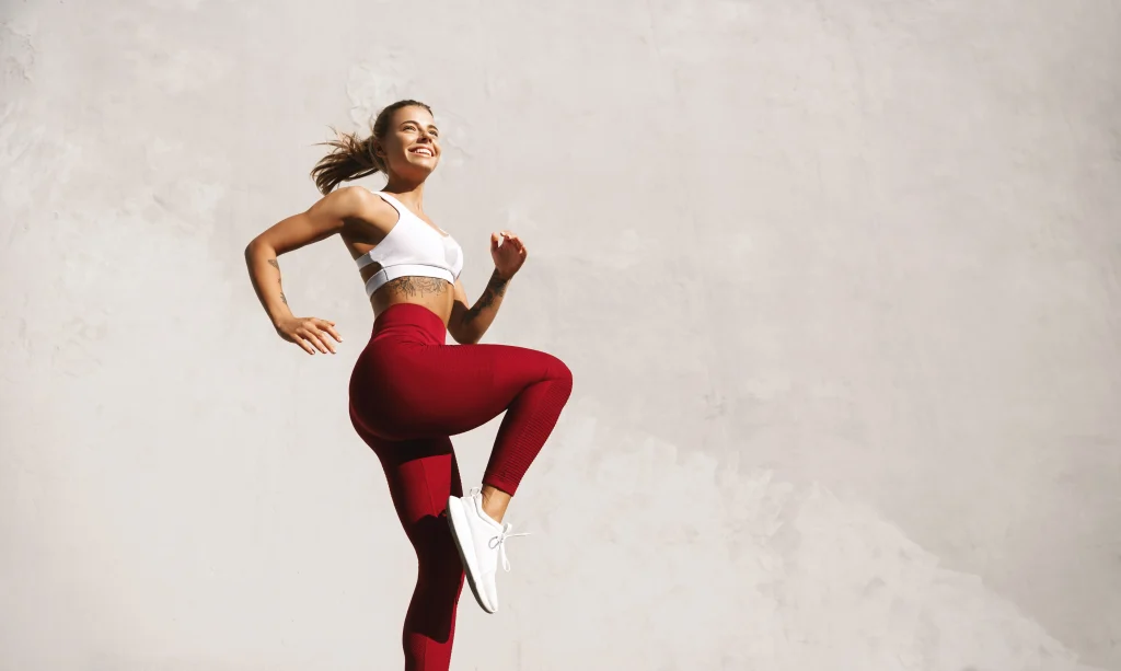 Person in athletic clothing performs a high-knee exercise against a light-colored wall outdoors.