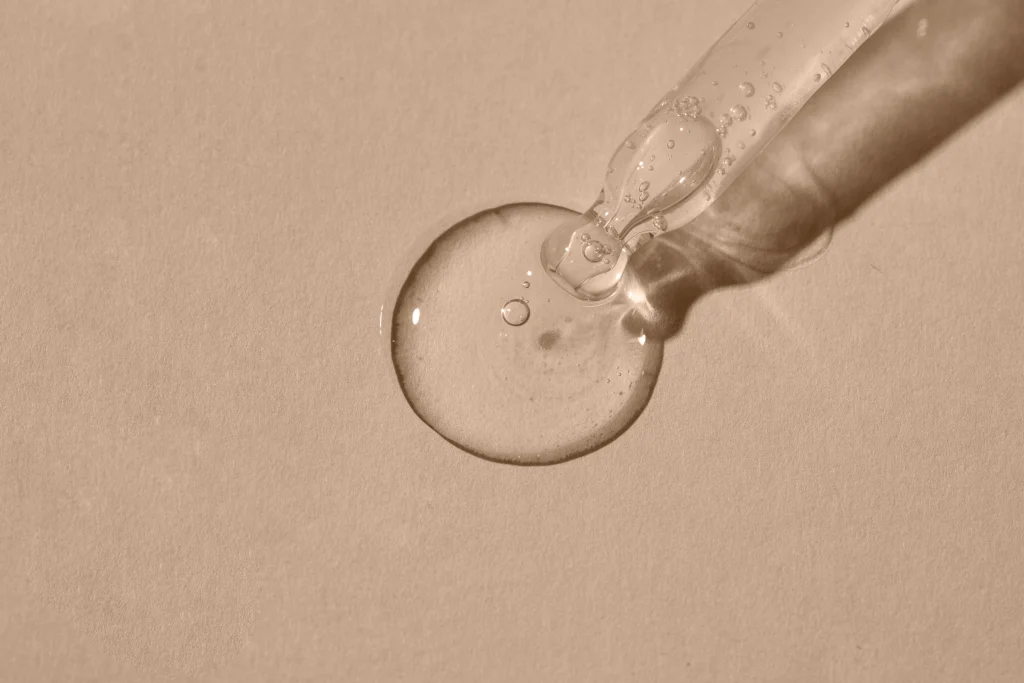 Dropper dispenses a clear skincare serum onto a smooth beige surface.