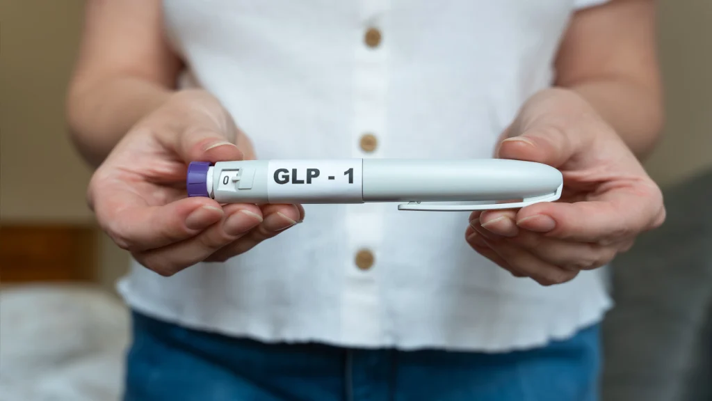 Person holds a GLP-1 injection pen with both hands against a blurred indoor background.