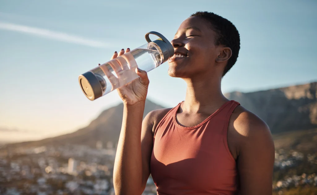 Person drinks water from a reusable bottle outdoors with mountains and sky in the background.