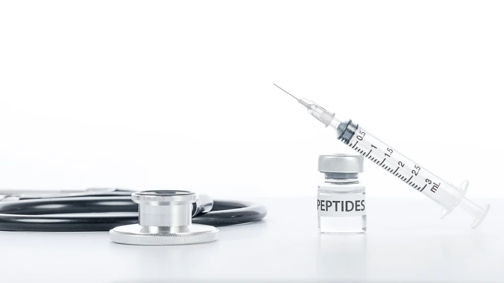 Syringe rests against a labeled peptide vial beside a stethoscope on a white medical surface