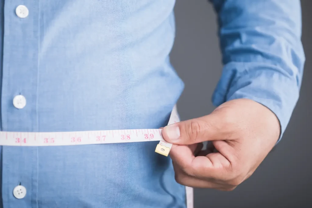 Close-up of a person measuring their waist with a measuring tape over a blue button-up shirt.